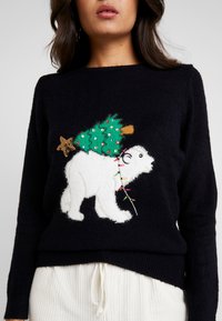 Donna che indossa un maglione nero con un orso bianco che porta un albero di Natale decorato, avvolto in luci colorate.