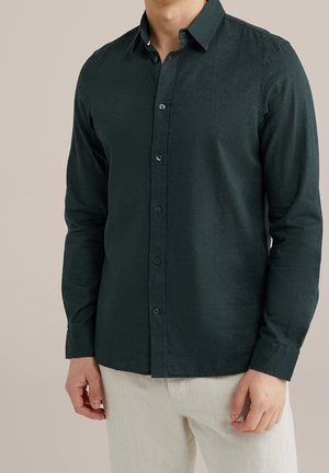 Homme portant une chemise boutonnée à manches longues vert foncé avec un pantalon beige, debout devant un fond clair uni.