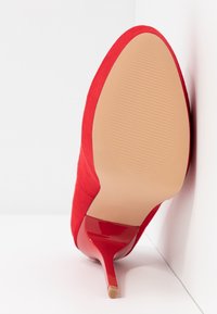 Escarpin en daim rouge à talon haut avec un bout pointu et un accent rouge brillant sur le talon, présentant une semelle beige texturée.