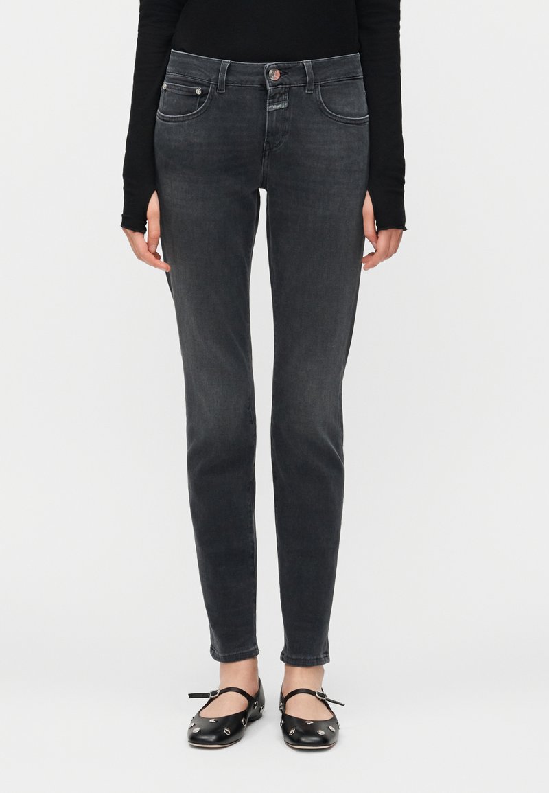 Femme portant un haut noir à manches longues, un jean skinny noir délavé, et des ballerines Mary Jane noires avec des clous argentés, debout devant un fond blanc.