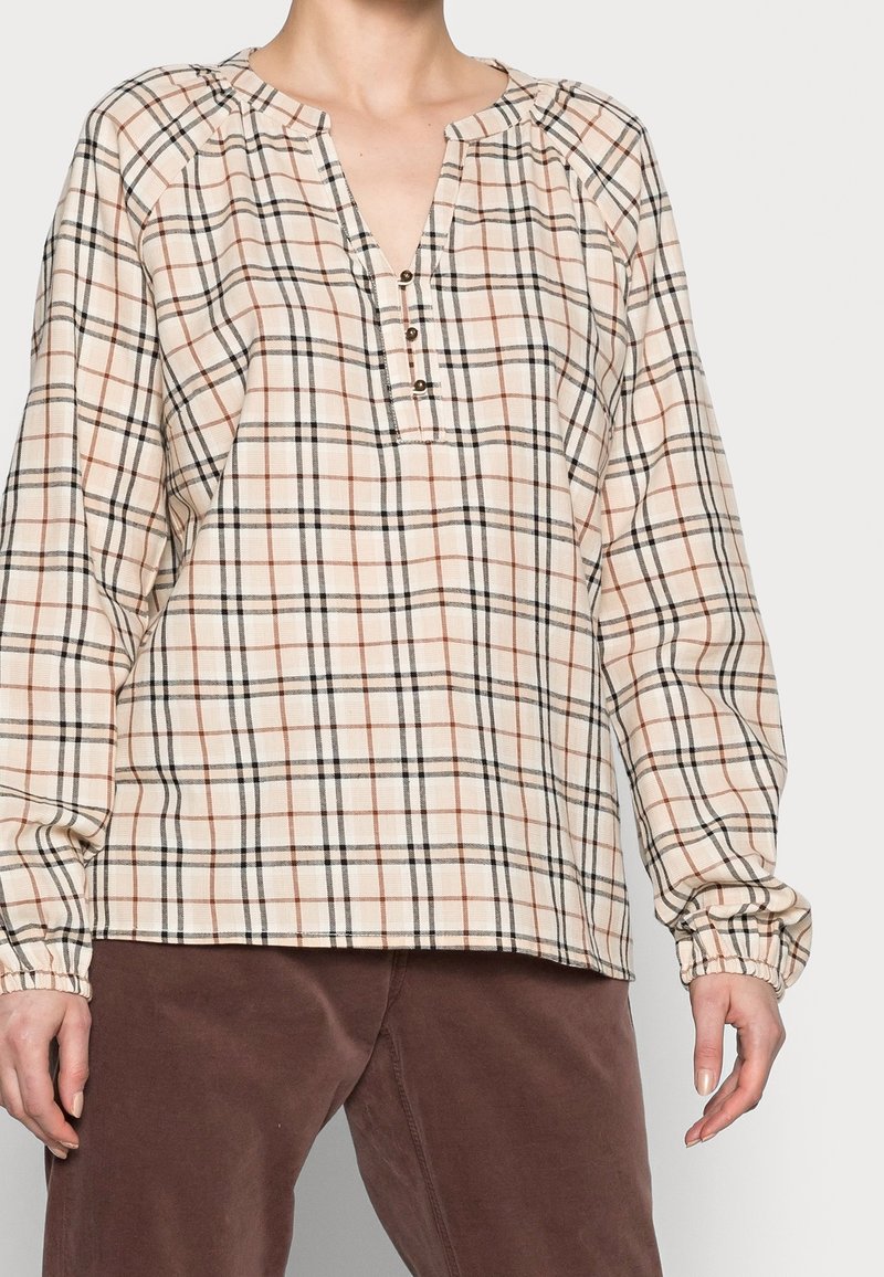 Personne portant un chemisier beige à carreaux manches longues avec un col boutonné et un pantalon marron sur un fond clair uni.