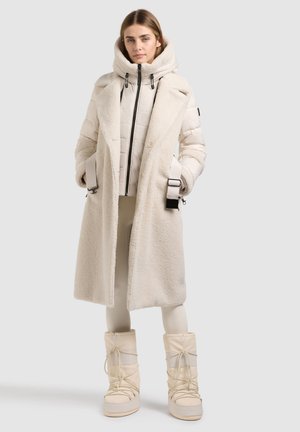 Jeune femme portant un long manteau teddy couleur crème, une veste matelassée zippée, des leggings et des bottes d'hiver lacées, debout devant un fond uni.