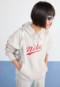 Gaiši pelēks kapuce ar sarkaniem akcentiem un lielu sarkanu Nike logo. Tam ir kapuce ar auklu un priekšējā kabata. Valkāts ar sudraba aksesuāriem.