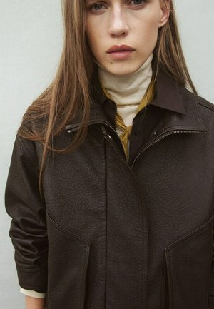 Giovane donna con capelli lunghi indossa una giacca di pelle scura e testurizzata sopra una maglietta nera e un dolcevita beige, in piedi davanti a uno sfondo neutro.