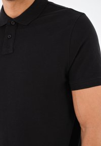 Polo noir en tissu texturé. Doté d'une patte de boutonnage à trois boutons et de manches courtes avec des poignets côtelés. Pas de motifs ou d'accents visibles.