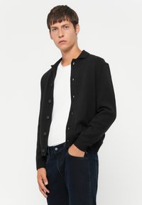 Svart strikket cardigan med krage, med knappelukking foran, ribbestrikkede mansjetter og strukturert stoff, brukt over en hvit t-skjorte.