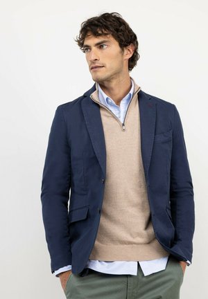Hombre con pelo castaño rizado que lleva un blazer azul marino, un jersey beige con cremallera hasta el pecho, una camisa azul clara y pantalones verdes, de pie con las manos en los bolsillos.