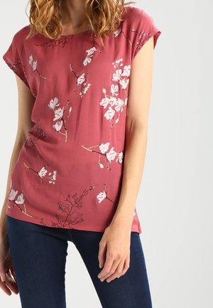 Žena v krátkém růžovém topu s bílým květinovým vzorem a tmavě modrých džínách stojí na jednobarevném světlém pozadí.