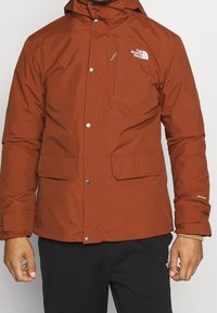 Hnědá zateplená bunda se zipem a patentkami na přední straně, dvěma předními kapsami a logem na hrudi, vybavená odnímatelnou kapucí a nastavitelnými manžetami.