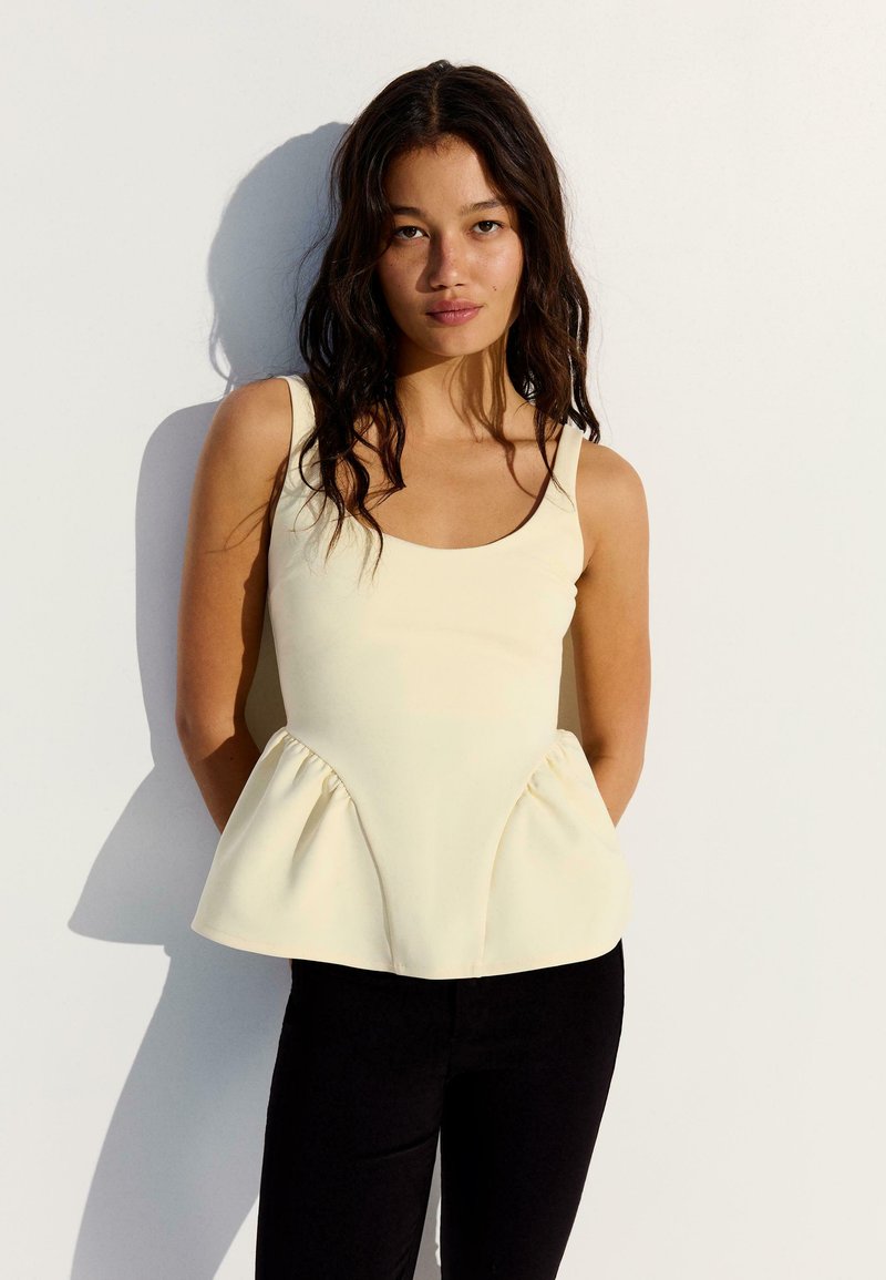 Jeune femme aux longs cheveux foncés portant un haut peplum crème sans manches et un pantalon noir, debout contre un mur blanc avec une ombre.