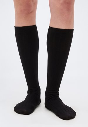 Jambes portant des chaussettes noires côtelées montantes jusqu'aux genoux, debout sur une surface blanche avec les pieds légèrement écartés.