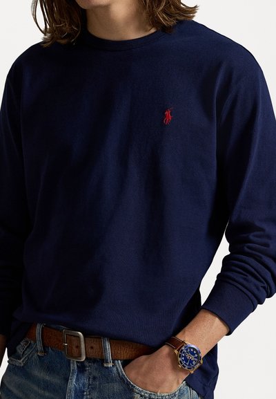 Marineblauwe longsleeve t-shirt met geribbelde ronde hals, voorzien van een klein rood logo op de borst. Gekocht met lichtgewassen jeans en een bruine riem.