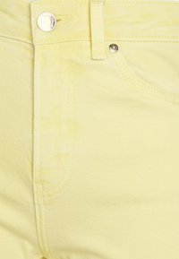 Tecido de denim amarelo claro, apresentando um fecho com botões na parte da frente, costuras trapadas e um contorno de bolso visível com uma textura suave.