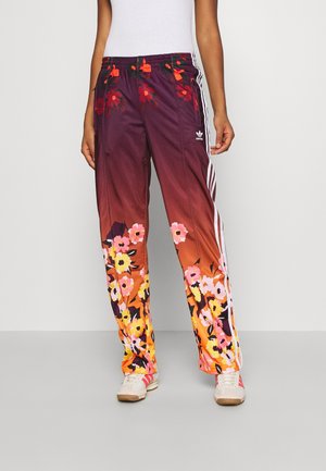 Person iført blomsterfarvede Adidas træningsbukser med hvide striber på siderne og hvide sneakers mod en ensfarvet baggrund.
