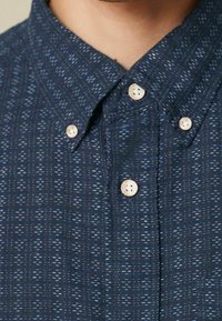 Camicia blu navy con bottoni, caratterizzata da un tessuto testurizzato con un sottile motivo a macchie, e bottoni bianchi lungo la patta e il colletto.