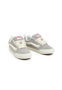 Sneaker in suede grigio con lacci bianchi, dettaglio a striscia nera e toppa con logo rosso. Il design presenta una silhouette low-top e accenti testurizzati.