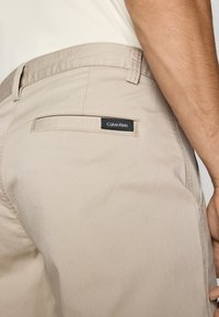 Béžové bavlnené nohavice s zadnou vreckou a čiernou etiketou Calvin Klein. Dizajn zahŕňa hladkú textúru a strih prispôsobený na mieru.