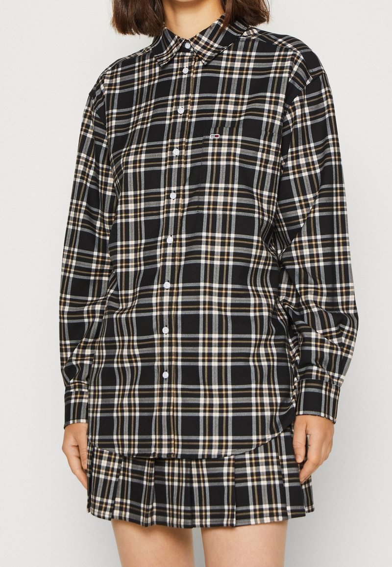 Camicia a quadri nera e marrone con bottoni, maniche lunghe, colletto a punta e tasca sul petto, abbinata a una gonna nera plissettata.