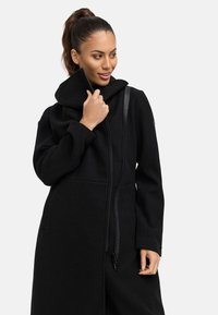 Manteau en laine noir avec un col haut, fermeture éclair et poches latérales. Il présente une finition texturée et des détails de zip contrastants.