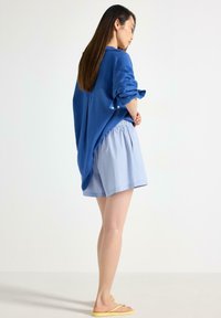 Blå oversized skjorte med oprullede ærmer, parret med lyseblå stribet shorts og gule strandflip-flops; glat tekstur og afslappet pasform.