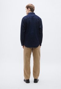 Μπλε σκούρο overshirt από τζιν με μυτερό γιακά, μακριά μανίκια και επίπεδο τελείωμα· σετ με παντελόνι καφέ κορντόρι και μαύρα παπούτσια.