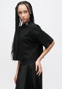 Μαύρο κοντό t-shirt με στρογγυλή λαιμόκοψη και κοντά μανίκια, με το λογότυπο "HUGO" στο ύψος του ώμου. Το υλικό φαίνεται λείο.