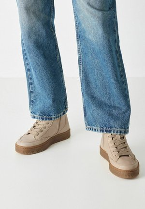 Beige high-top sneakers with textured sides, round toe, and a brown rubber sole. Paired with light blue unhemmed jeans.