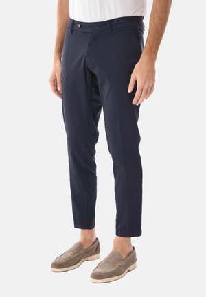 Uomo che indossa pantaloni aderenti blu navy e scarpe beige senza lacci, in piedi contro uno sfondo chiaro uniforme.