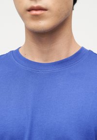 Νεαρό άτομο που φοράει απλό μπλε t-shirt με στρογγυλή λαιμόκοψη, εμφανίζοντας τον λαιμό, το πηγούνι και το άνω μέρος του στήθους πάνω σε ουδέτερο φόντο.