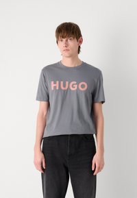 Pilkas marškinėliai su apvaliu kaklu, ant kurių užrašyta "HUGO" koralinės rožinės spalvos. Audinys atrodo lygus. Modelis dėvi juodas džinsus.