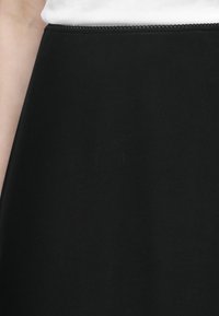Jupe noire taille haute avec une ceinture à bord festonné, portée avec un haut blanc, montrée de la taille jusqu'à mi-cuisse.
