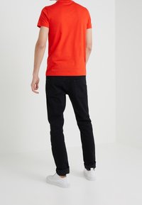 Camisa de tirantes roja combinada con jeans ajustados negros y zapatillas blancas, mostrando un look casual desde la parte posterior.