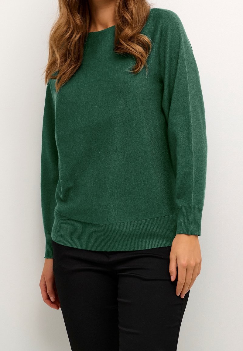Femme portant un pull vert uni à manches longues et un pantalon noir, debout devant un fond neutre uni.