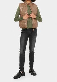 Brązowa pikowana kamizelka z wysokim kołnierzem, noszona na zielonej koszuli na guziki, w zestawieniu z czarnymi dżinsami i błyszczącymi czarnymi butami.