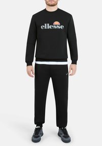 Felpa nera con logo "ellesse" in grigio e rosso, abbinata a pantaloni jogger neri. Presenta polsini a costine e vita elastica. Sneakers nere.