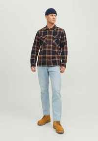 Camisa de franela a cuadros en azul oscuro, verde y rojo, con dos bolsillos en el pecho; combinada con vaqueros azul claro y botas de trabajo marrones.