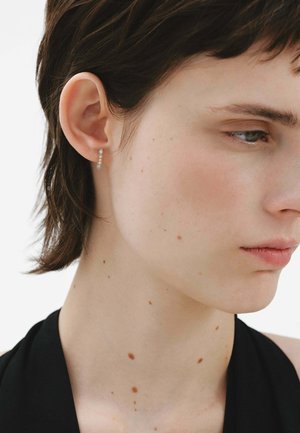 Pendiente en tono dorado con un diseño vertical que presenta pequeñas piedras preciosas transparentes, en contraste con un tono de piel claro y cabello corto oscuro.