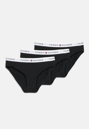 Три черни слипове Tommy Hilfiger с бели ластици на кръста, върху които е изписано името на марката и логото, разположени един върху друг на бял фон.