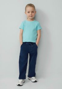 Lichtblauw geribd T-shirt met korte mouwen en marineblauwe joggingbroek met trekkoord. Witte sneakers met grijze accenten. Kind dat staat met de handen in de zakken.