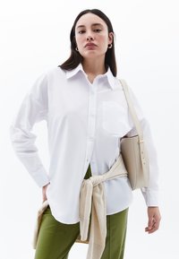 Camicia bianca con bottoni, maniche lunghe, tasca sul lato sinistro e vestibilità comoda, abbinata a pantaloni verdi e una borsa beige a tracolla.