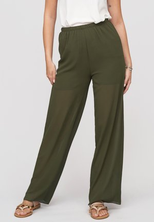 Femme portant un pantalon large vert olive avec taille élastique et des sandales beiges, debout devant un fond uni.