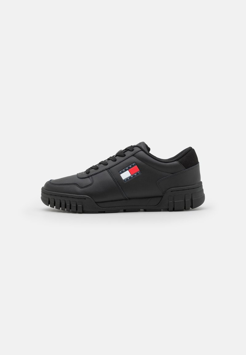 Baskets basses noires avec une semelle épaisse et des lacets, avec un petit logo rectangulaire à rayures rouges, blanches et bleues sur le côté.