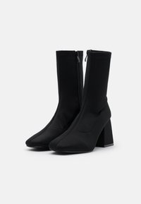 Botines negros hasta el tobillo con un upper de textil suave, punta afilada y tacón cuadrado. Presenta una cremallera lateral y un detalle de costura visible.