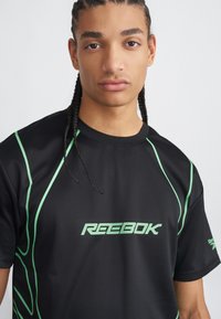 Čierne atletické tričko má zelené akcenty a logo "Reebok" na prednej strane. Vyrobené z ľahkej látky s okrúhlym výstrihom.
