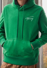 Felpa verde con una consistenza morbida, dotata di cappuccio, tasca frontale, polsini a coste e logo bianco sul petto.