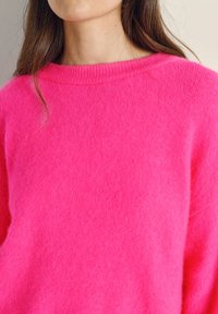 Donna che indossa un maglione rosa acceso peloso con scollo tondo, che mostra parte del collo e dei capelli castani.