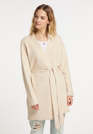 Frau trägt einen cremefarbenen Cardigan mit Gürtel über einem weißen Top und zerrissene hellblaue Jeans, schaut leicht zur Seite.