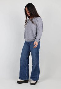 Un pull gris surdimensionné, léger et à coupe décontractée, associé à un jean flare bleu foncé. Les chaussures sont blanches avec des semelles noires. Fond neutre.