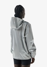 Persona con el pelo trenzado, vistiendo una chaqueta con capucha plateada brillante y pantalones cortos negros, vista desde atrás sobre un fondo liso.