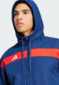 Marineblå hoodie med en rød vandret stribe, justerbar snøre i hætten og sølvlogo detaljer på brystet og skulderen. Glat stofstruktur.